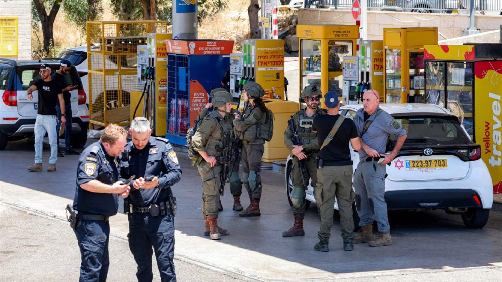 Security forces in tactical gear stand in a street area with a white car and yellow kiosks behind them, appearing to supervise a checkpoint.