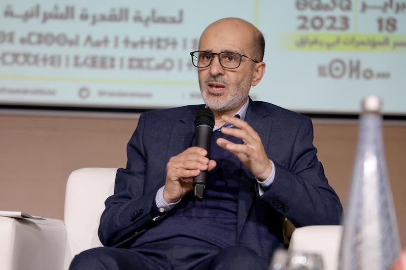 Man wearing glasses and a dark suit speaks into a handheld microphone while seated on a white chair at a panel discussion.