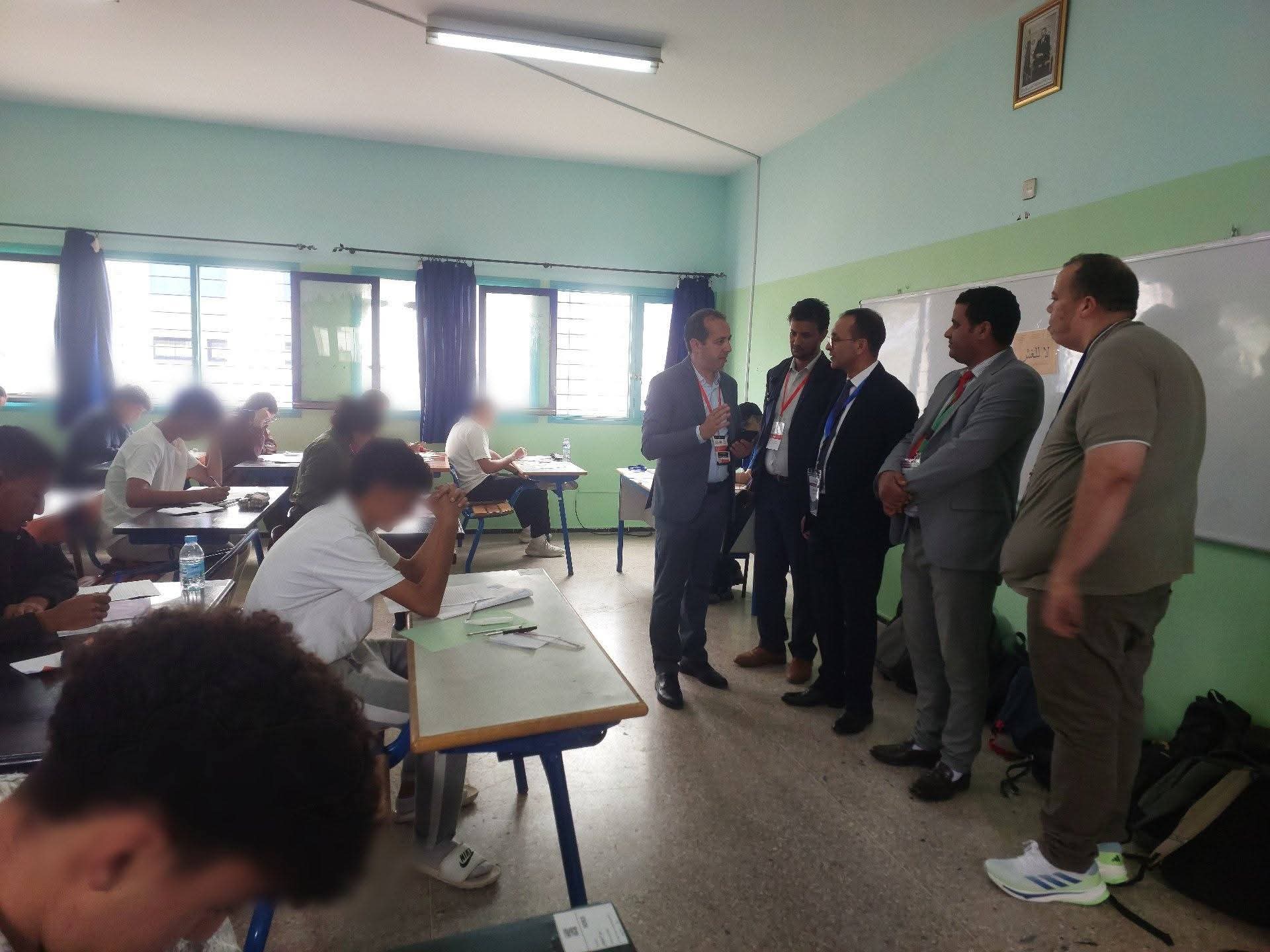 FB_IMG_1780334799033 | MCG24 | موقع إلكتروني مغربي شامل متجدد على مدار الساعة Classroom scene with students at desks on the left and five men in suits standing and talking near the front of the room.