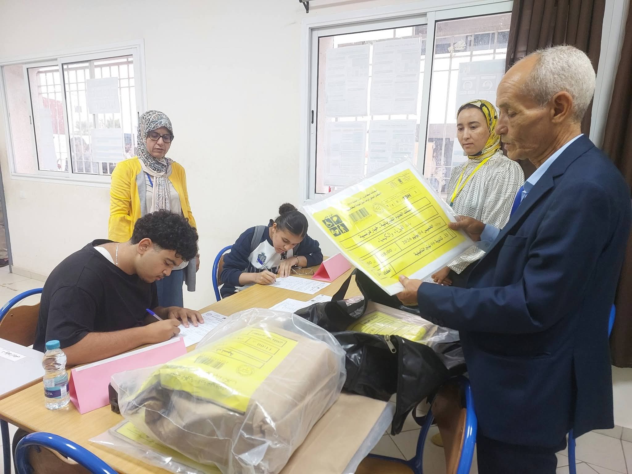 Man in a blue suit holds a large yellow form while two women wearing hijabs assist and others write at a table in a bright room with pink folders nearby.