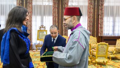 FB_IMG_1780944029226 | MCG24 | موقع إلكتروني مغربي شامل متجدد على مدار الساعة Smiling woman in blue and black attire receives a green item from a man in gray ceremonial robes and a red fez during a formal ceremony.