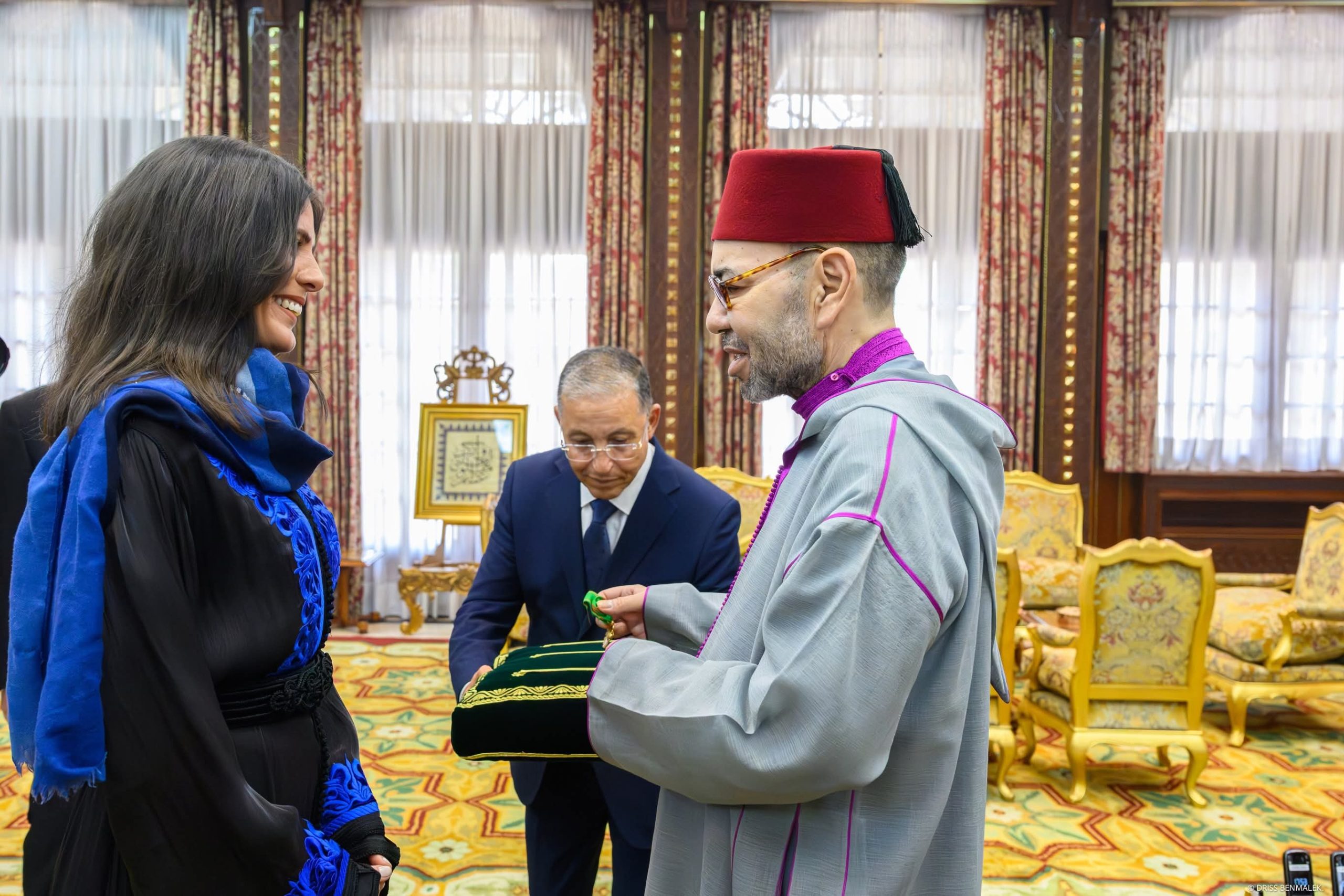 Smiling woman in blue and black attire receives a green item from a man in gray ceremonial robes and a red fez during a formal ceremony.