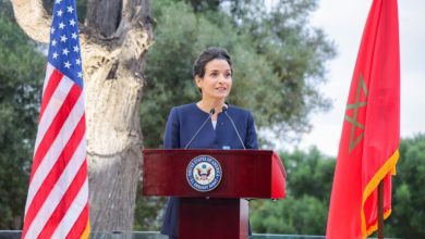 A woman in a navy suit gives a formal speech at a wooden podium outdoors, flanked by the U.S. flag on one side and a red flag on the other.