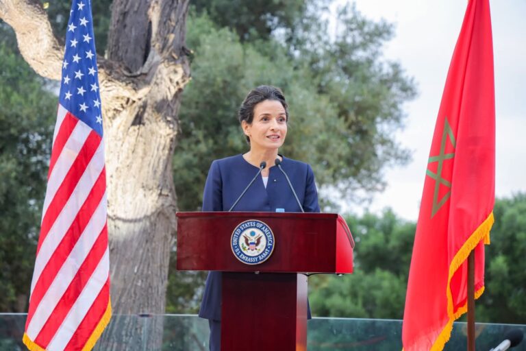 A woman in a navy suit gives a formal speech at a wooden podium outdoors, flanked by the U.S. flag on one side and a red flag on the other.