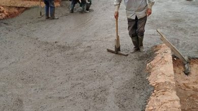 FB_IMG_1781174664402 | MCG24 | موقع إلكتروني مغربي شامل متجدد على مدار الساعة Workers lay fresh concrete on a rural road, using shovels and rakes beside a stone wall and white building at dusk/dawn light.