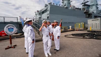 Sailors in white dress uniforms standing on a ship's deck with a large gray warship in the background.”] ,