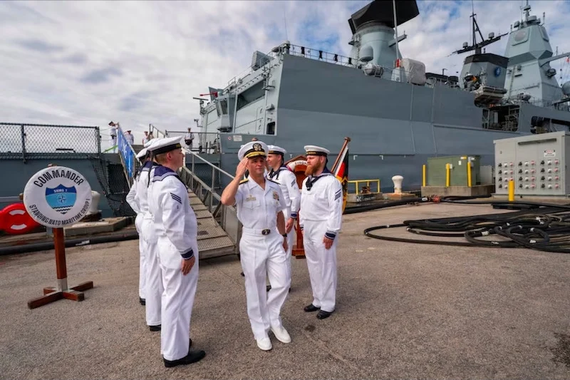 Sailors in white dress uniforms standing on a ship's deck with a large gray warship in the background.”] ,