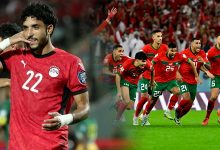 Soccer player in a red jersey with the number 22 saluting, while teammates celebrate on the field in the background.