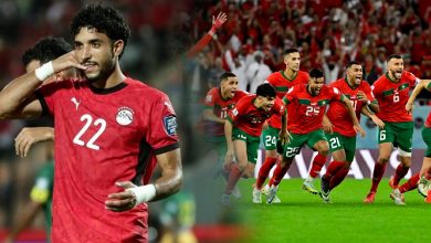 Soccer player in a red jersey with the number 22 saluting, while teammates celebrate on the field in the background.