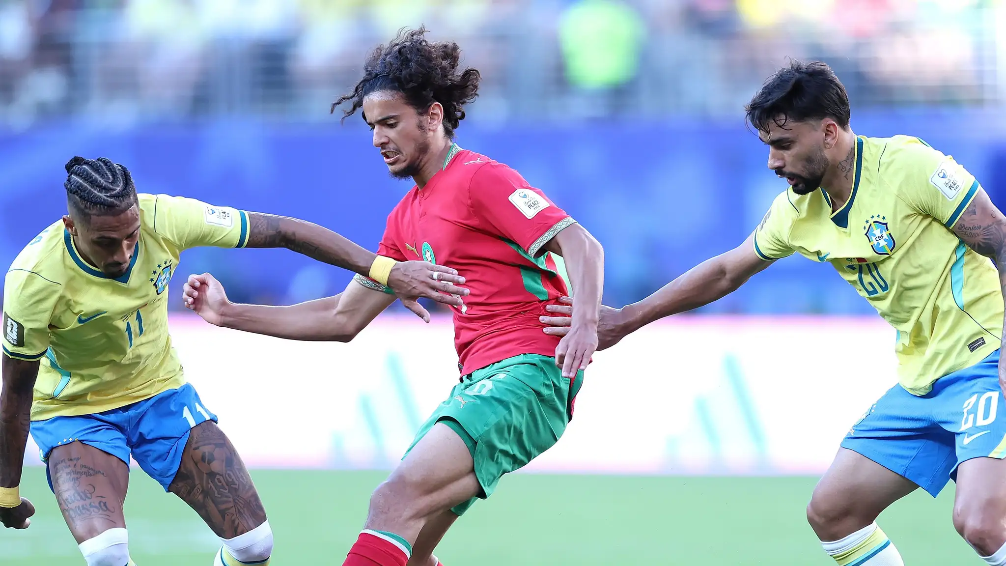 GettyImages-2281751288jpg | MCG24 | موقع إلكتروني مغربي شامل متجدد على مدار الساعة Three soccer players clash for the ball: two in yellow Brazil kits flank a player in red and green on a green field.