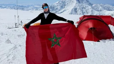 Person standing in a snowy mountain camp, holding a Moroccan flag with a red tent in the background, ski poles nearby.