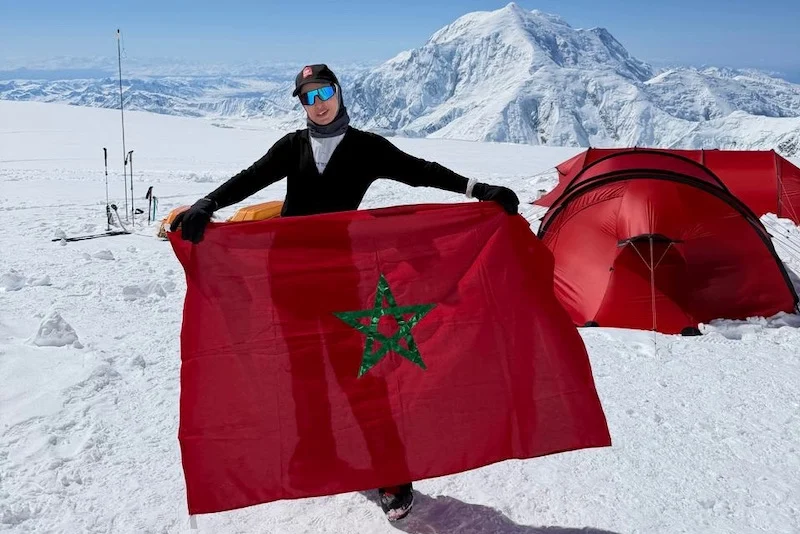 Hind-Zemmama | MCG24 | موقع إلكتروني مغربي شامل متجدد على مدار الساعة Person standing in a snowy mountain camp, holding a Moroccan flag with a red tent in the background, ski poles nearby.