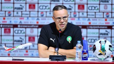 IMG-20260317-WA0024-1536x852 | MCG24 | موقع إلكتروني مغربي شامل متجدد على مدار الساعة Athletic coach in a black polo with Moroccan crest speaks at a press conference, microphone in foreground and sponsor backdrop behind him.