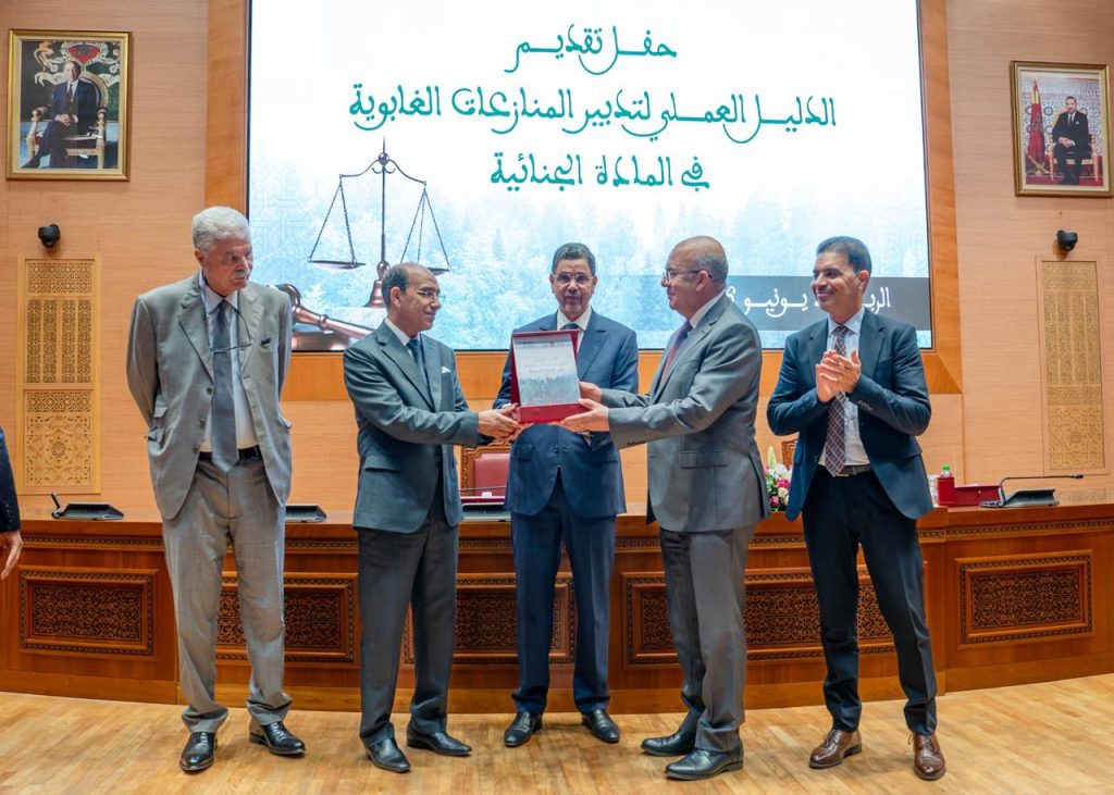 Award ceremony: two men in suits exchange a red-framed plaque as others applaud on a wooden stage with a large Arabic text backdrop and portrait-filled walls behind them.