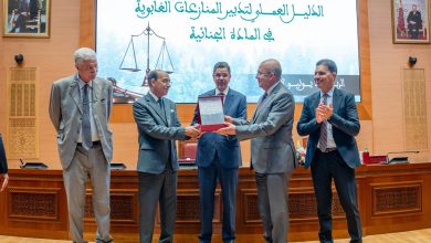 Award ceremony: two men in suits exchange a red-framed plaque as others applaud on a wooden stage with a large Arabic text backdrop and portrait-filled walls behind them.