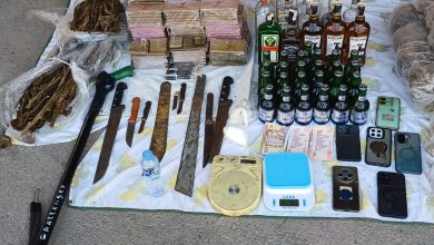 Display of contraband on a tarp: stacked boxes, various knives and blades, a scale, cash, and many liquor bottles arranged for display.