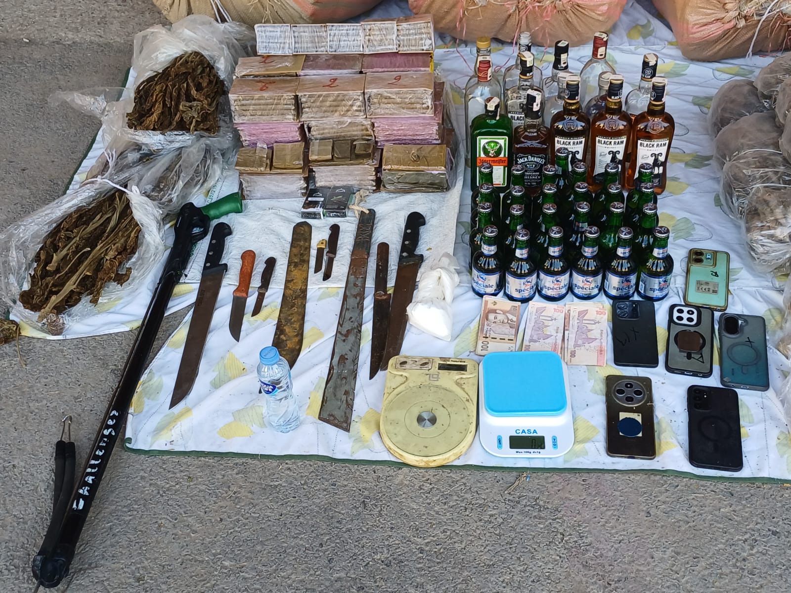 Display of contraband on a tarp: stacked boxes, various knives and blades, a scale, cash, and many liquor bottles arranged for display.