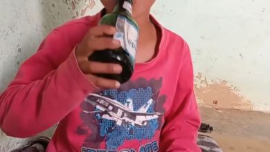 Young child in a pink shirt sits cross‑legged, drinking from a bottle, with eyes covered by a gray bar against a peeling wall background.