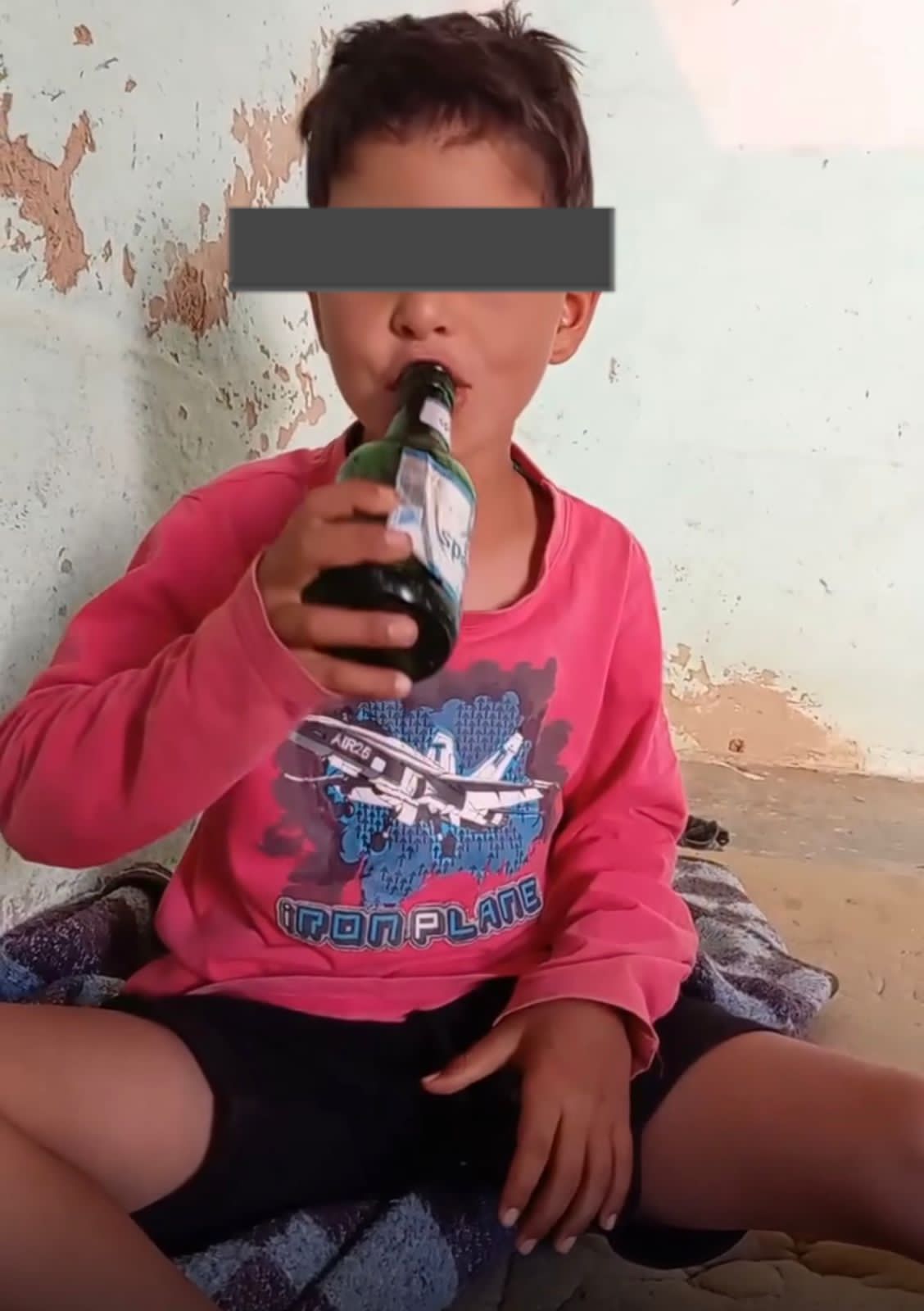 IMG-20260607-WA0000 | MCG24 | موقع إلكتروني مغربي شامل متجدد على مدار الساعة Young child in a pink shirt sits cross‑legged, drinking from a bottle, with eyes covered by a gray bar against a peeling wall background.