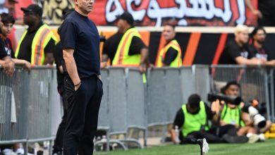 وهبي | MCG24 | موقع إلكتروني مغربي شامل متجدد على مدار الساعة Man in a navy shirt and white sneakers stands on a grassy sports field, with security staff and a crowd behind a red banner in the stands.