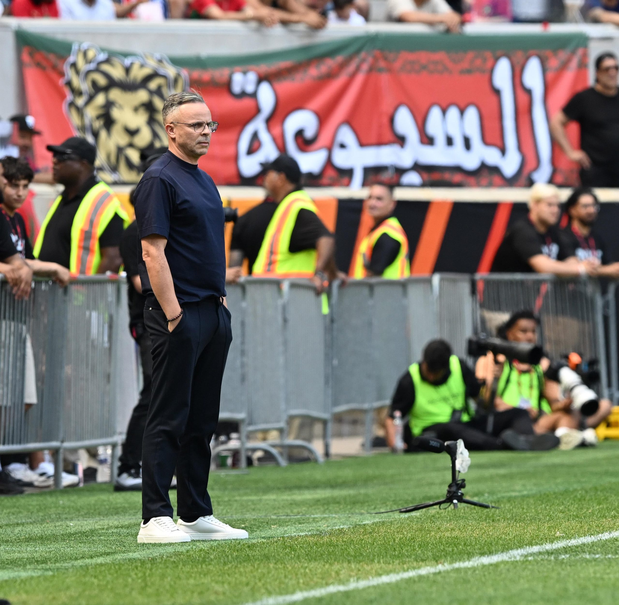 وهبي | MCG24 | موقع إلكتروني مغربي شامل متجدد على مدار الساعة Man in a navy shirt and white sneakers stands on a grassy sports field, with security staff and a crowd behind a red banner in the stands.