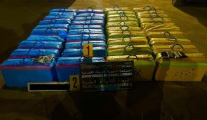 Rows of blue and green rectangular packages with handles laid out as police evidence, with a sign reading'Police Technique et Scientifique' in front.