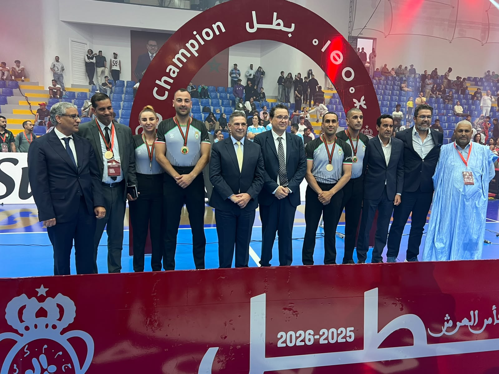 Group of medalists and officials posing on a sports court in front of a large red Champion arch and cheering crowd.