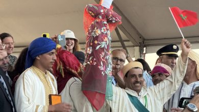 Group of Moroccan performers in traditional dress celebrating, one person holds up a colorful floral shawl-wrapped figure atop a stick amid smiling crowd and a red flag in the background