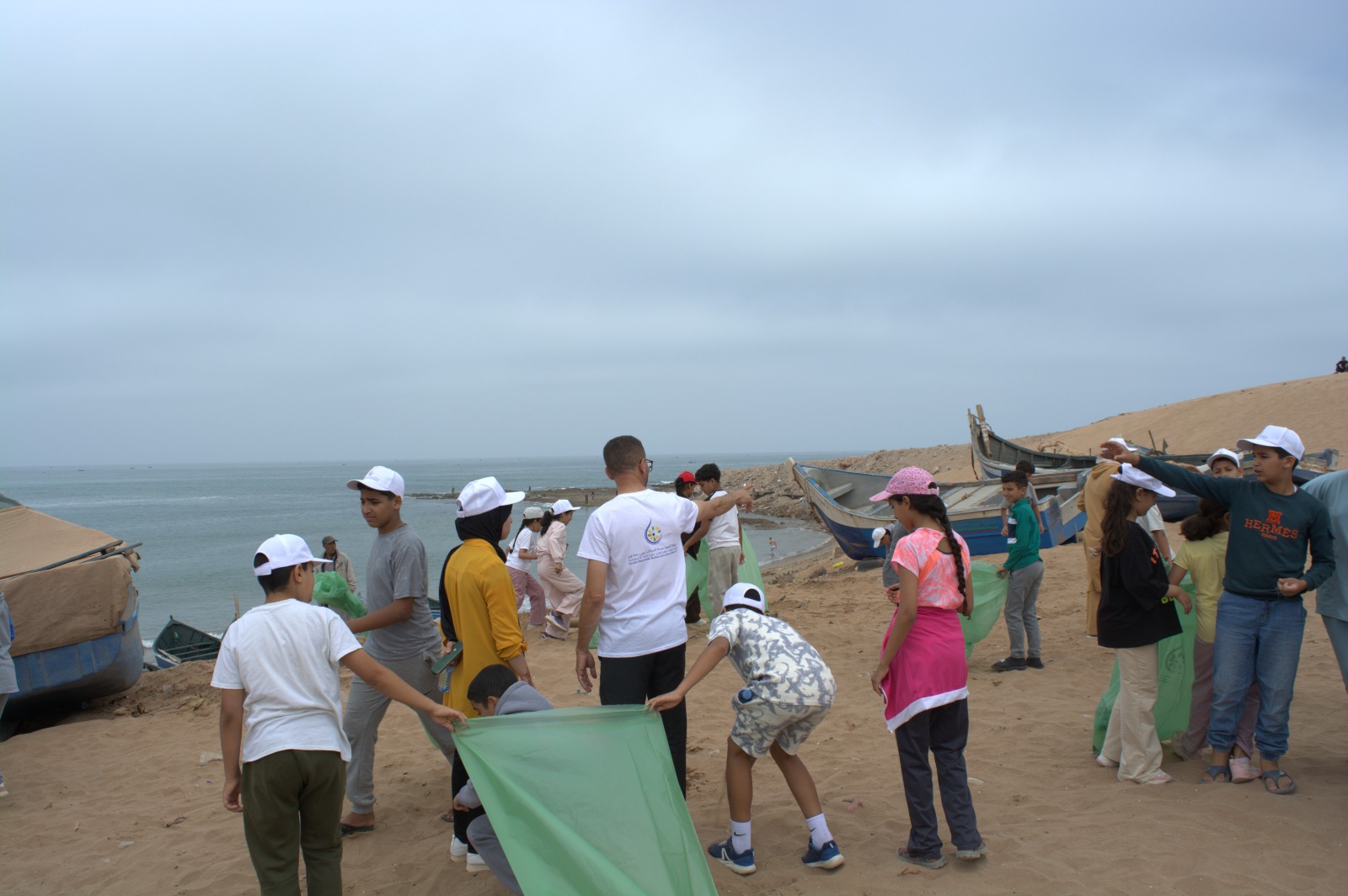 IMG-20260616-WA0005 | MCG24 | موقع إلكتروني مغربي شامل متجدد على مدار الساعة Children on a sandy beach collect litter into green bags; boats sit at the shore, ocean in the background.",
