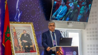IMG-20260616-WA0079 | MCG24 | موقع إلكتروني مغربي شامل متجدد على مدار الساعة Man in a blue suit giving a lecture at a podium with a laptop and microphones, a large framed portrait and Moroccan flag beside him.