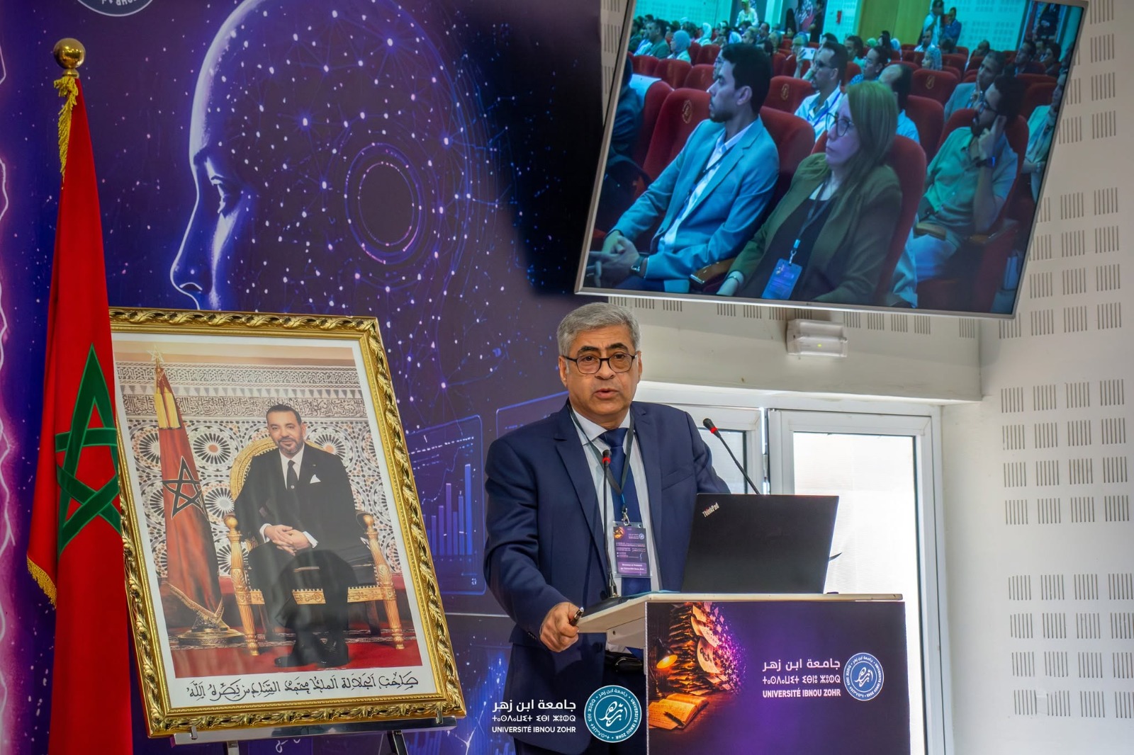 IMG-20260616-WA0079 | MCG24 | موقع إلكتروني مغربي شامل متجدد على مدار الساعة Man in a blue suit giving a lecture at a podium with a laptop and microphones, a large framed portrait and Moroccan flag beside him.