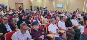 Diverse audience seated in red theater-style chairs listening to a talk in a modern conference hall.