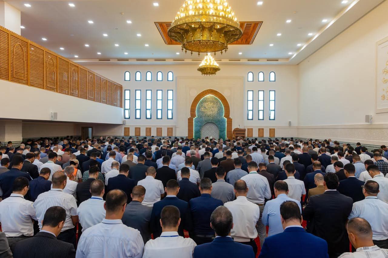 IMG-20260619-WA02252 | MCG24 | موقع إلكتروني مغربي شامل متجدد على مدار الساعة Large crowd of men praying in a mosque hall, facing an ornate turquoise-tiled mihrab with arches and gold chandeliers overhead.