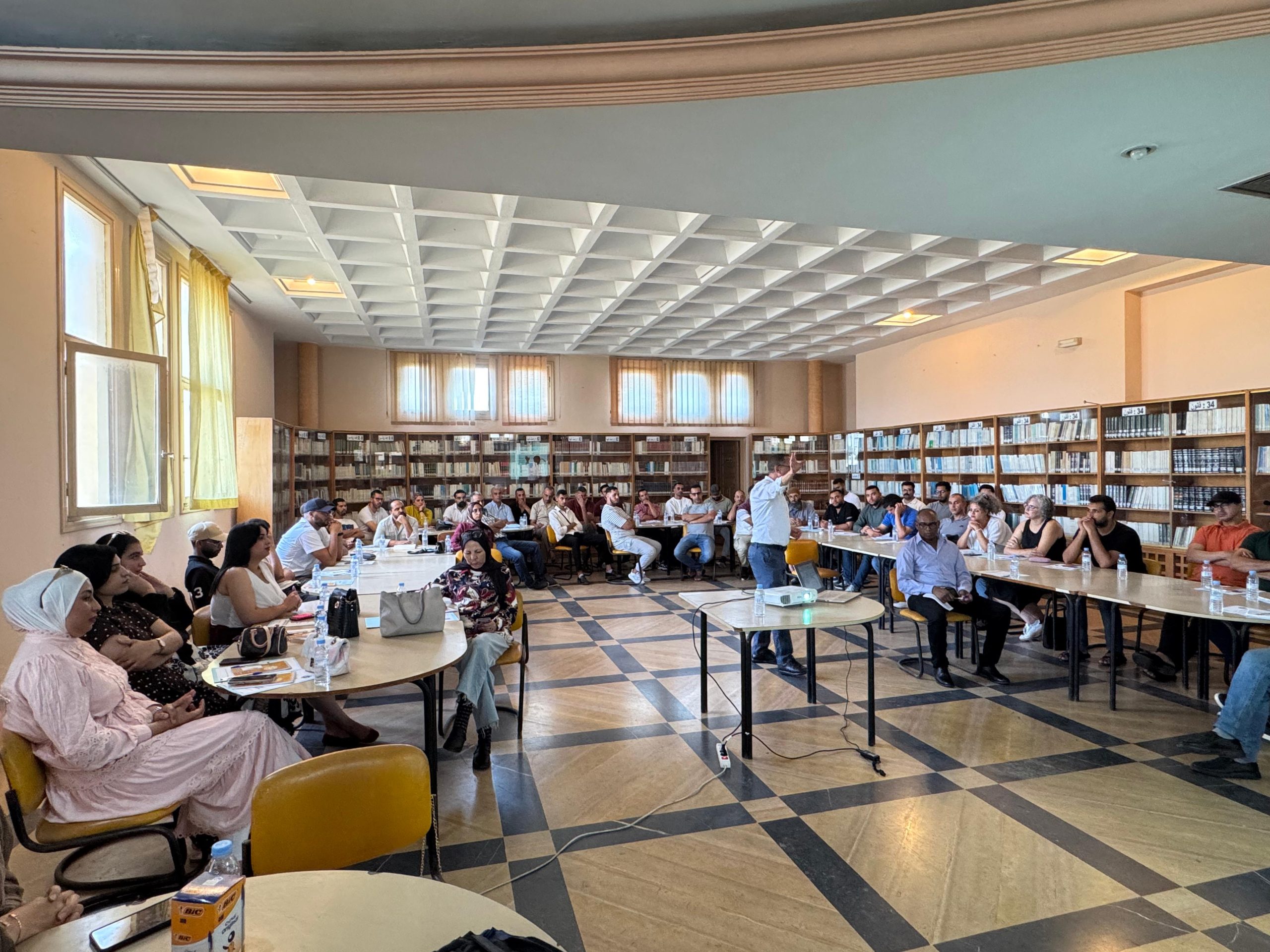 IMG-20260620-WA0092 | MCG24 | موقع إلكتروني مغربي شامل متجدد على مدار الساعة Group of people seated around tables in a library, listening to a presenter standing at the front.