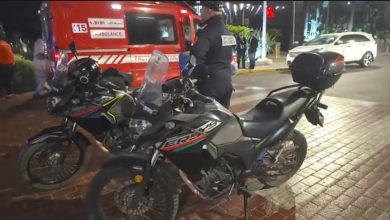 IMG-20260621-WA0053 | MCG24 | موقع إلكتروني مغربي شامل متجدد على مدار الساعة Nighttime street scene with two motorcycles parked on a brick sidewalk; an ambulance and people in the background suggest an emergency or incident.