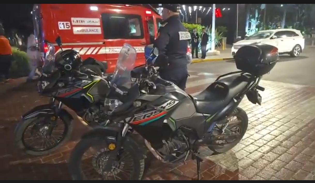 IMG-20260621-WA0053 | MCG24 | موقع إلكتروني مغربي شامل متجدد على مدار الساعة Nighttime street scene with two motorcycles parked on a brick sidewalk; an ambulance and people in the background suggest an emergency or incident.
