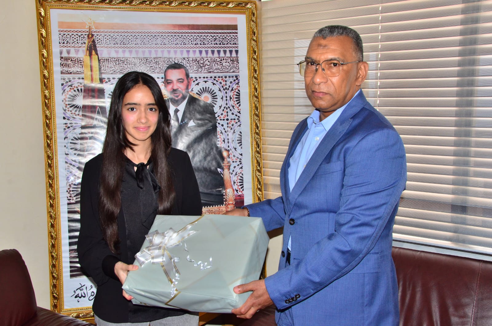 IMG-20260621-WA0191 | MCG24 | موقع إلكتروني مغربي شامل متجدد على مدار الساعة Girl receiving a wrapped gift from a man in a blue suit in a living-room/office setting with a framed poster in the background.
