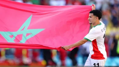 IMG_0111 | MCG24 | موقع إلكتروني مغربي شامل متجدد على مدار الساعة Soccer player in white and red kit holding a large pink flag with a teal star design behind him in a stadium crowd