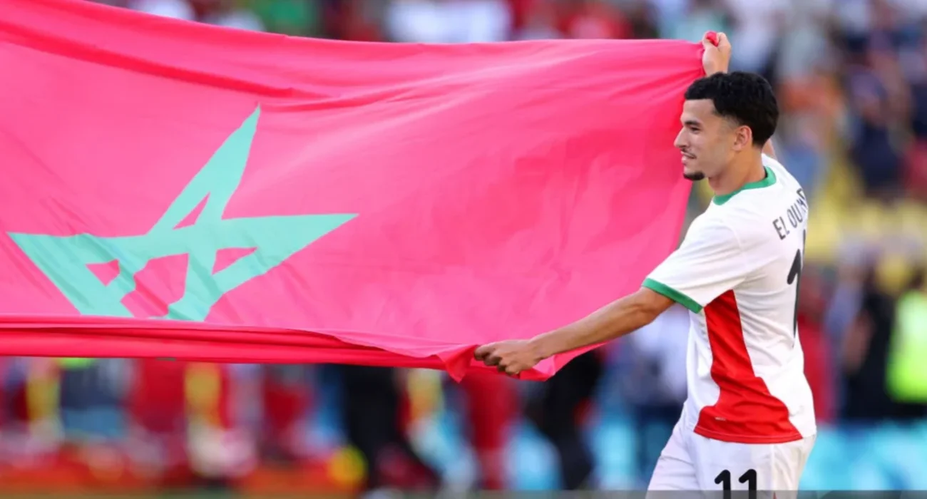 IMG_0111 | MCG24 | موقع إلكتروني مغربي شامل متجدد على مدار الساعة Soccer player in white and red kit holding a large pink flag with a teal star design behind him in a stadium crowd