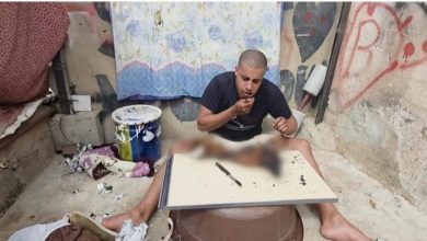 Man sits cross‑legged at a makeshift table in a dirty, graffiti‑covered room, with a blurred, injured body on the table in front of him.