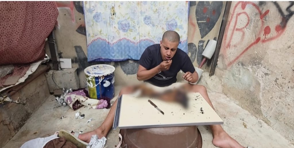 Man sits cross‑legged at a makeshift table in a dirty, graffiti‑covered room, with a blurred, injured body on the table in front of him.