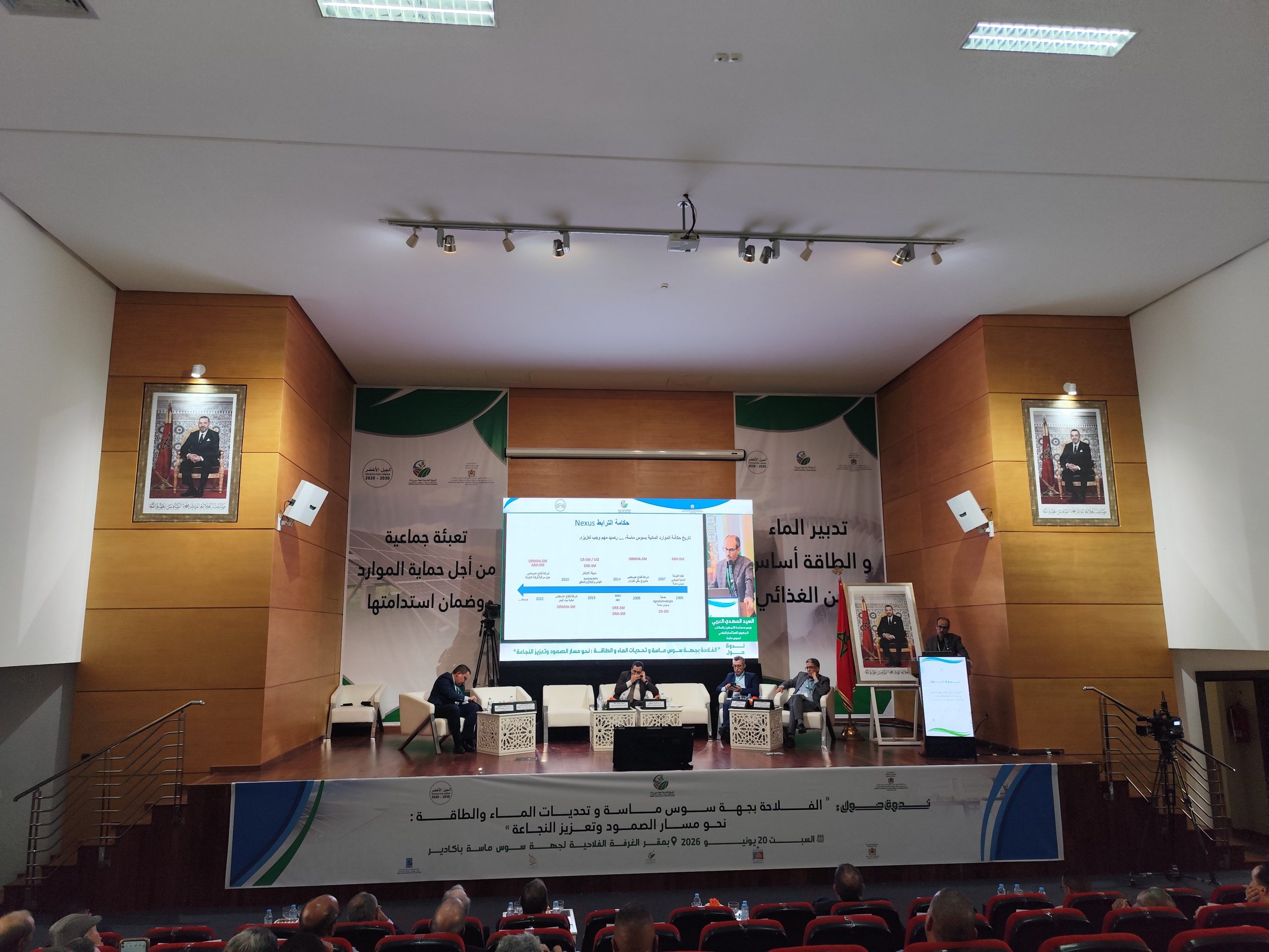 IMG_20260620_124419 | MCG24 | موقع إلكتروني مغربي شامل متجدد على مدار الساعة Conference stage with panelists seated at a long table, banners in Arabic behind them and portraits on either side of the walls.