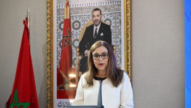 Woman wearing glasses speaks at a podium; Moroccan flag visible to her left and a framed portrait of King Mohammed VI behind her.