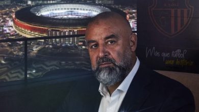 Portrait of a bearded man in a dark suit and white shirt on a terrace at night, with a brightly lit stadium behind him.