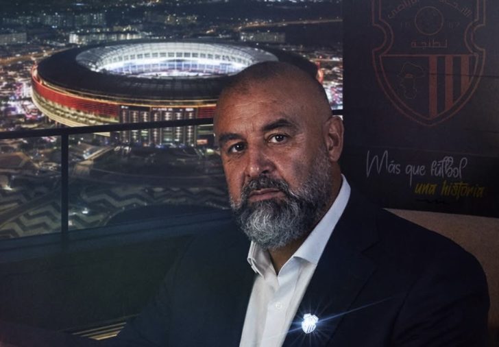 Portrait of a bearded man in a dark suit and white shirt on a terrace at night, with a brightly lit stadium behind him.