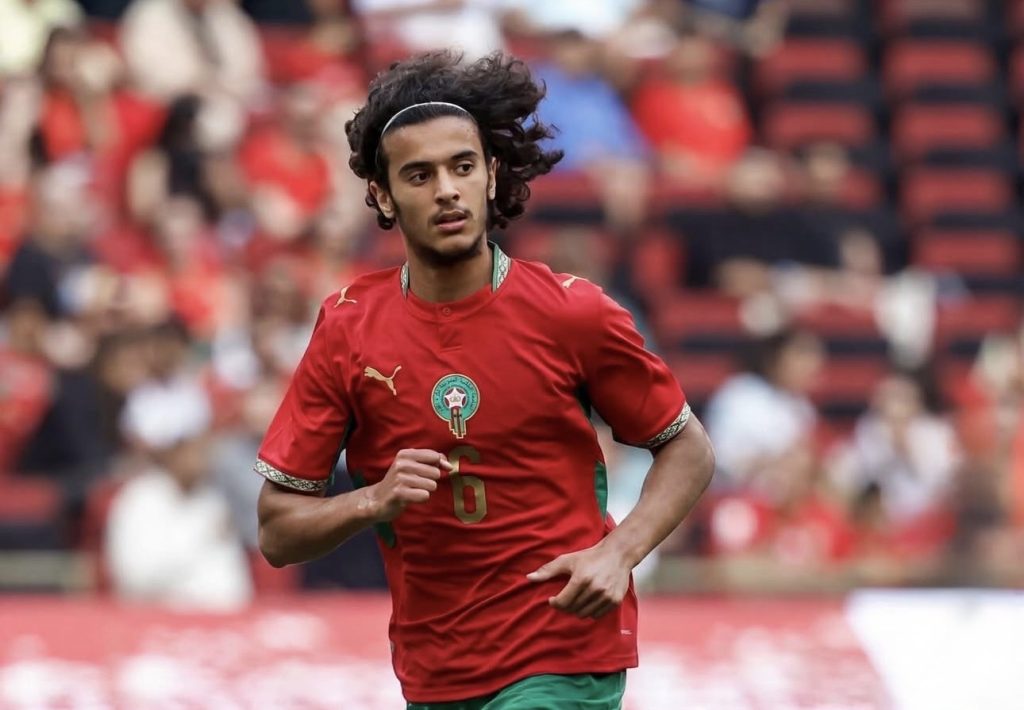 Soccer player in a red jersey with number 6, running on the field during a match, with blurred stadium crowd in the background.
