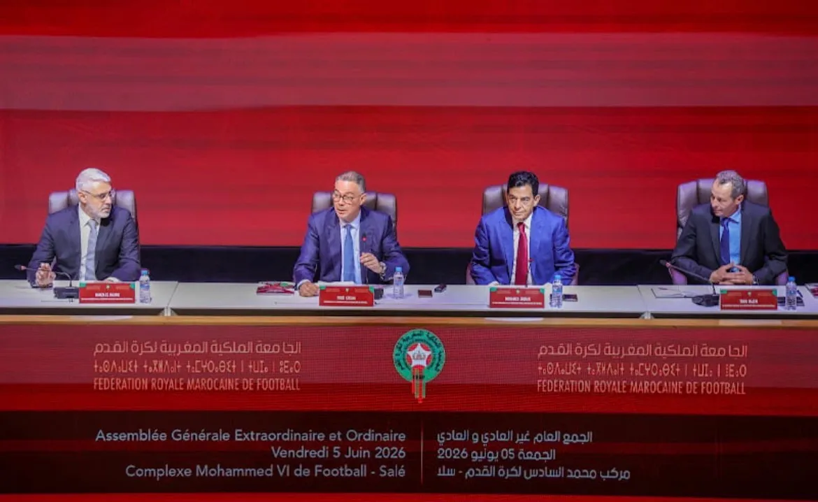 Four men in suits sit at a panel table on a stage, with a red backdrop and banners for the Moroccan Football Federation in Arabic and French behind them.