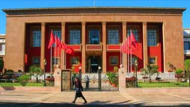 IMG_8953 | MCG24 | موقع إلكتروني مغربي شامل متجدد على مدار الساعة Front view of a red-brick government-style building with tall columns, flagpoles bearing red flags, and a gated entrance with a person walking by.