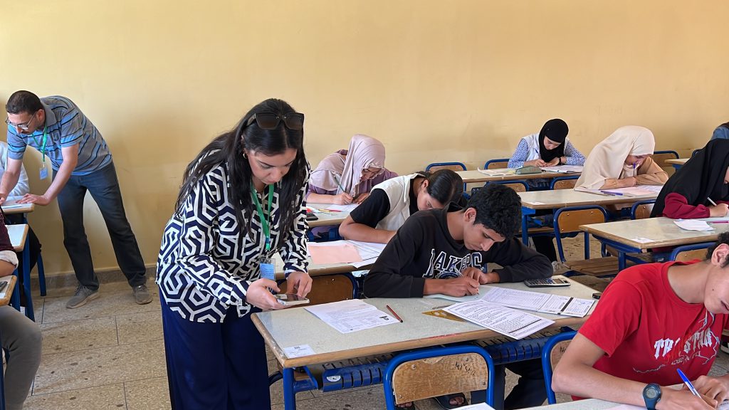 A classroom scene with students seated at desks taking a written exam while a teacher assists a student at the front left.