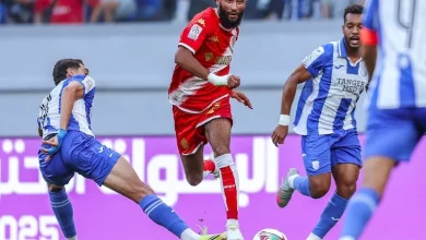 IRT-WAC1 | MCG24 | موقع إلكتروني مغربي شامل متجدد على مدار الساعة Soccer player in a red jersey leaps with the ball as blue-and-white defenders approach on a green pitch behind him, stadium spectators in the background.